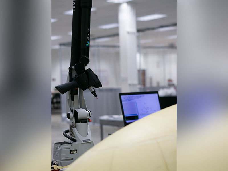 Image of the docked Romer Absolute Arm in a Rock West Composites manufacturing bay. It is located next to a piece of hardware and a computer screen.